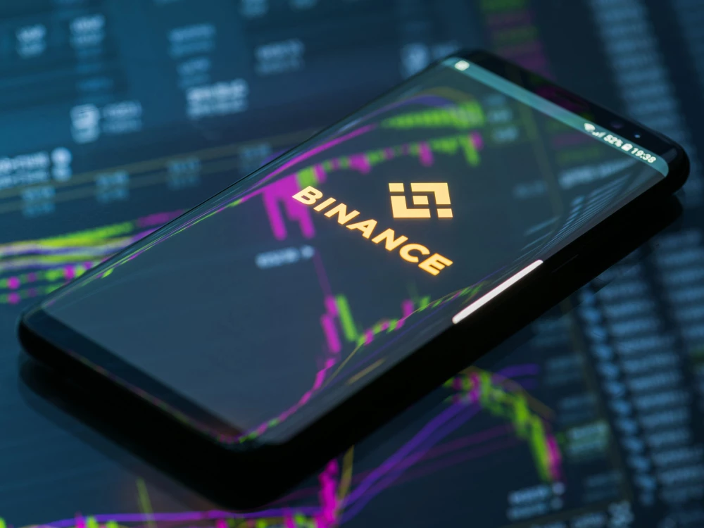 The Binance app open on a phone in front of crypto price charts.